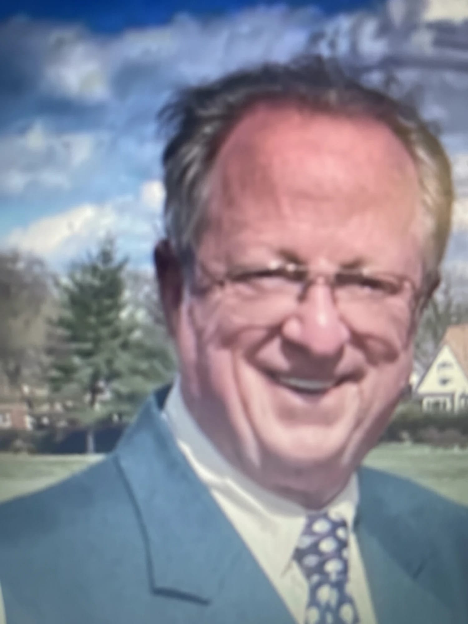 Dennis Riley, smiling, in a suit and patterned tie, outdoors on a sunny day.